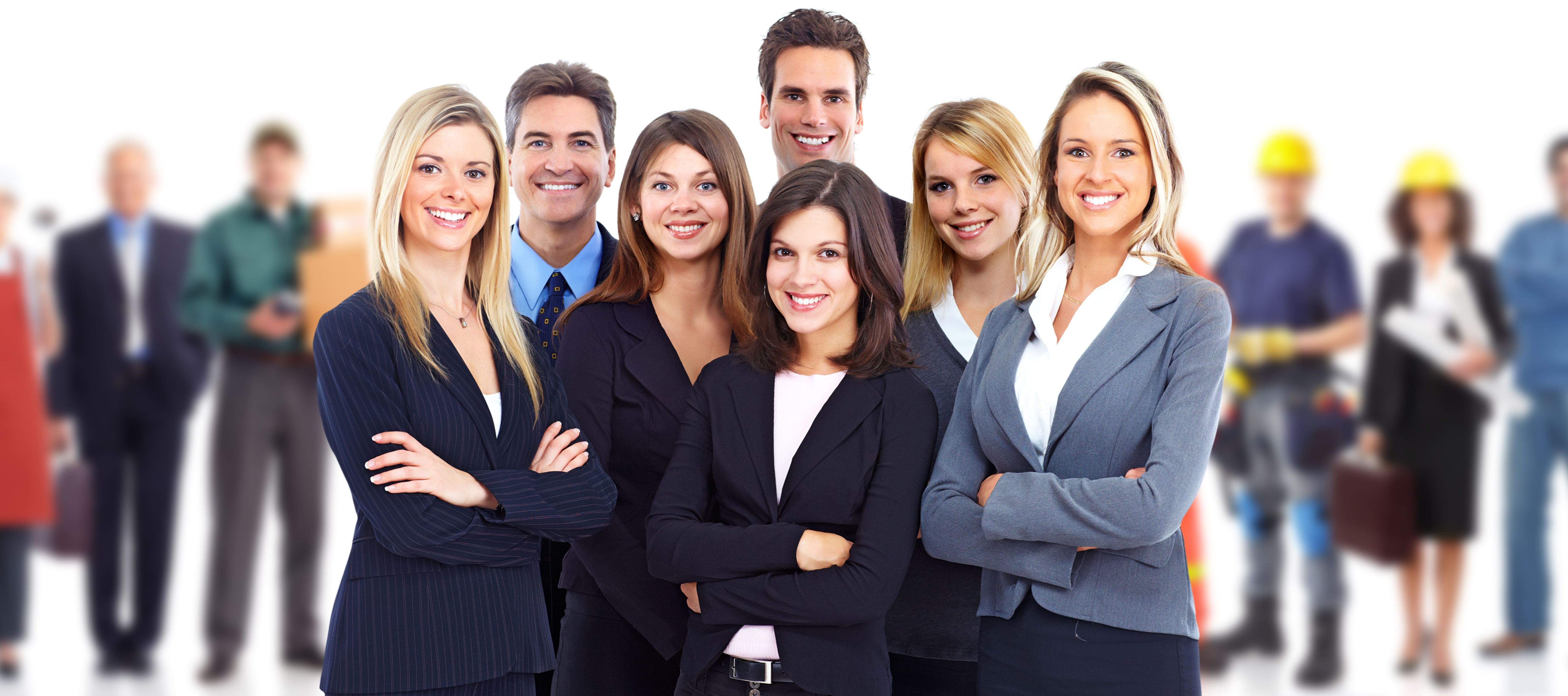 Das Bild zeigt eine Gruppe von Menschen. Die Personen im Vordergrund tragen Businesskleidung wie Anzüge und Blazer. Im Hintergrund sind weitere Menschen zu sehen, einige tragen Bauhelme und Arbeitsuniformen. Der Hintergrund ist leicht unscharf im Vergleich zum Vordergrund.